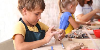 Young boy focused on sculpting clay with other children crafting in the background.