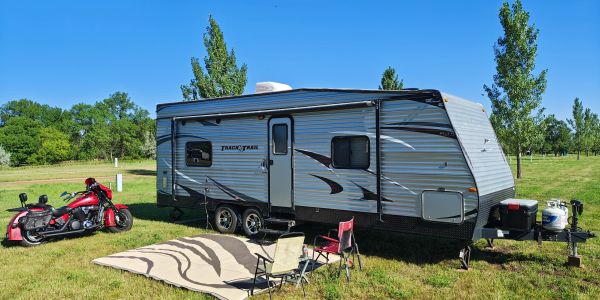 A travel trailer parked on grass with chairs and a motorcycle nearby.