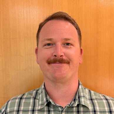 A man with a mustache wearing a green plaid shirt against a wooden background.