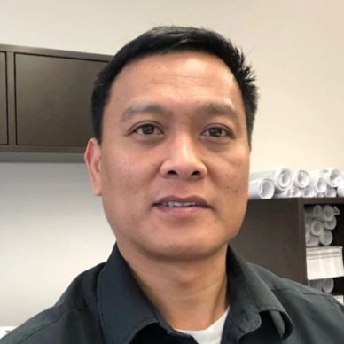 Man in black shirt posing indoors with rolled-up papers in background.