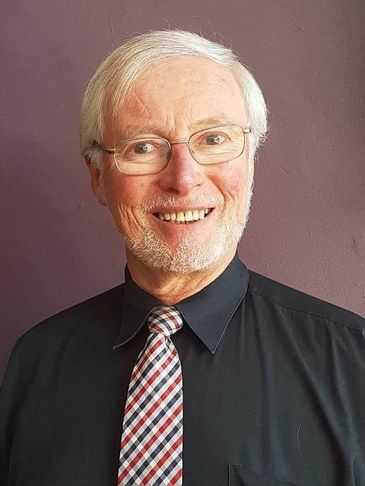Smiling elderly man with glasses and checkered tie.