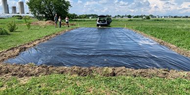 A black plastic tarp covers a rectangular area in a grassy field with people and a vehicle nearby.