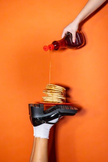 Syrup being poured over pancakes stacked on a sneaker against an orange background.