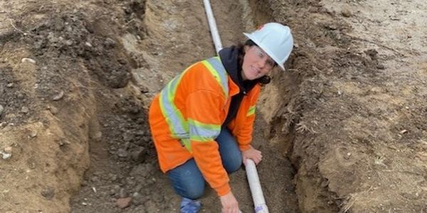 Worker in safety gear applying adhesive to a pipe in a trench at a construction site.
