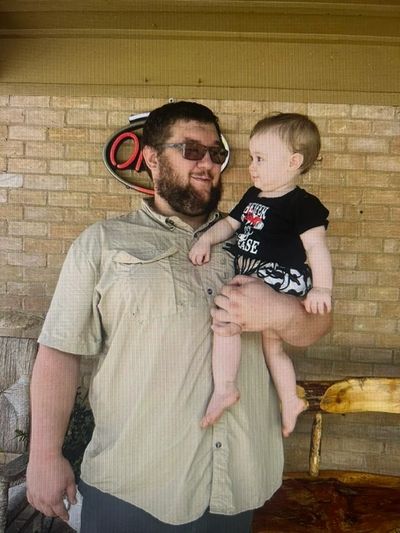 Man holding a baby, standing by a brick wall.