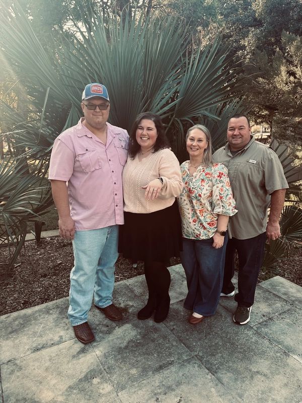 Four adults smiling together outdoors with greenery.