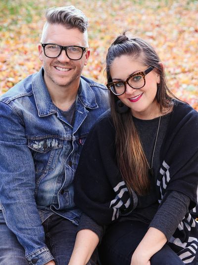 A smiling couple wearing glasses sitting outdoors in autumn leaves.