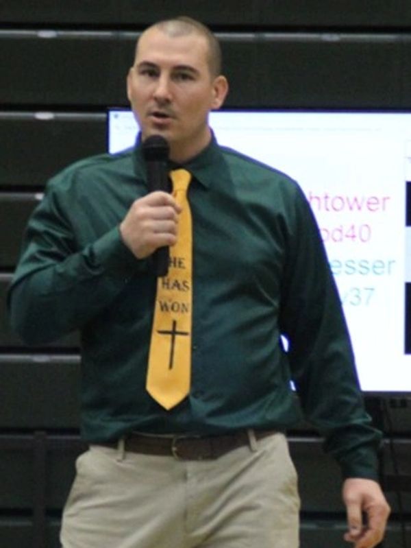 Man speaking into a microphone, wearing a green shirt and yellow tie with cross.