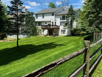 A white house with a wooden deck and a large green lawn enclosed by a rustic fence.