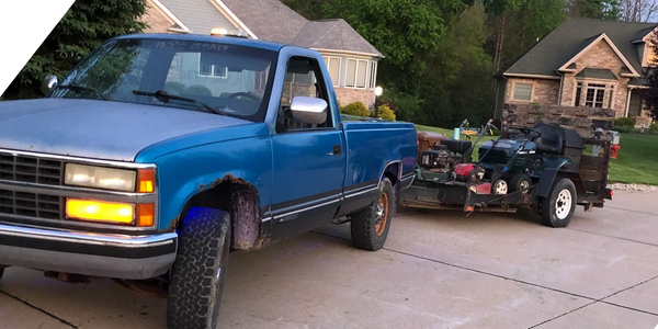 Old blue pickup truck towing a trailer with lawn equipment.