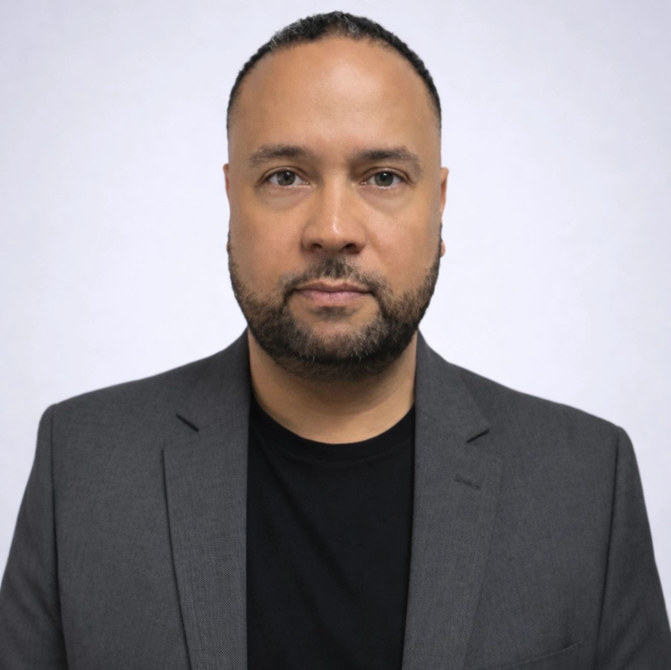 Man with a beard wearing a gray blazer and black shirt against a plain background.