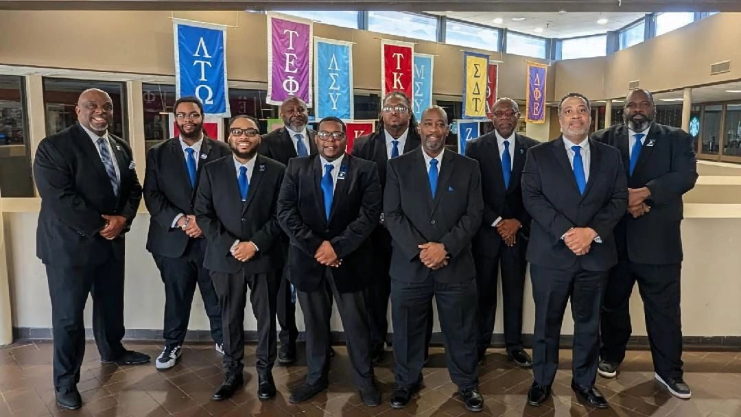 Group of men in suits with blue ties standing together indoors before fraternity banners.