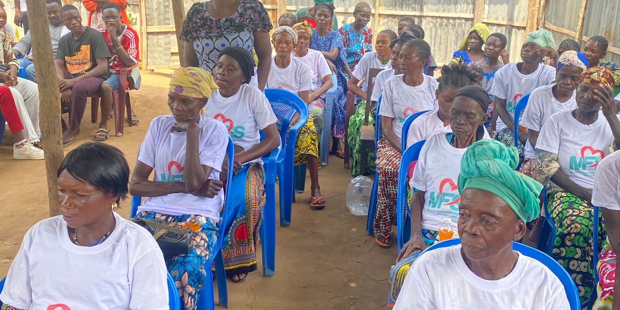 A group of elderly women seated in rows, wearing white shirts with colorful skirts and headwraps.