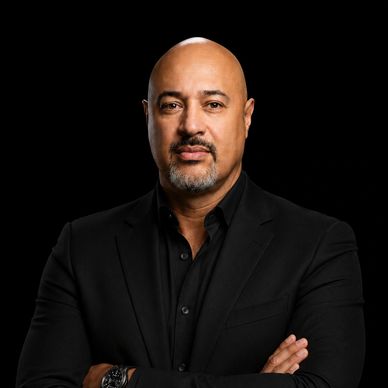 Confident man in a black suit with arms crossed against a black background.