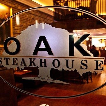 View through glass door with Oak Steakhouse logo into cozy restaurant interior.