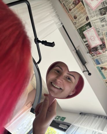 Person with red hair smiling at their reflection in a heart-shaped mirror.