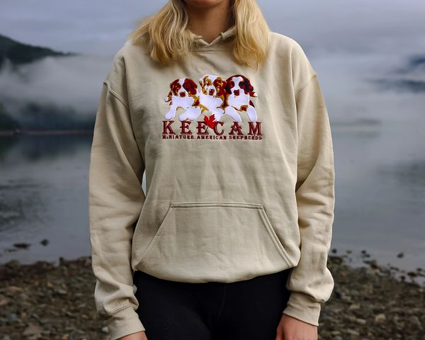Person wearing a beige hoodie with Miniature American Shepherd dog illustrations and text.