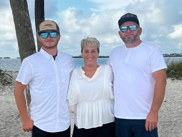 Three people standing together on a sandy beach with palm trees and water in the background.