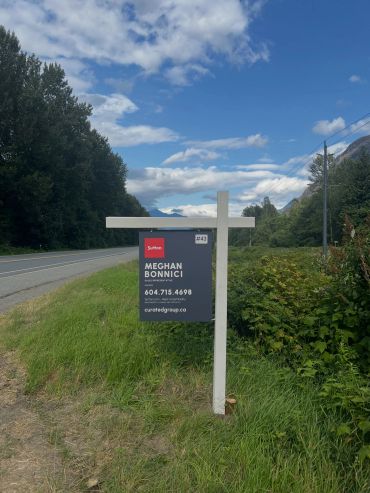 Real estate sign by a roadside with scenic mountains.