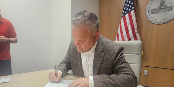 Man in a suit signing a document at a desk with an American flag behind him.
