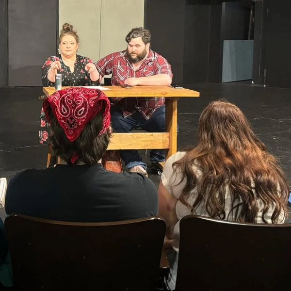 Two people sit at a table facing an audience in a theater setting.