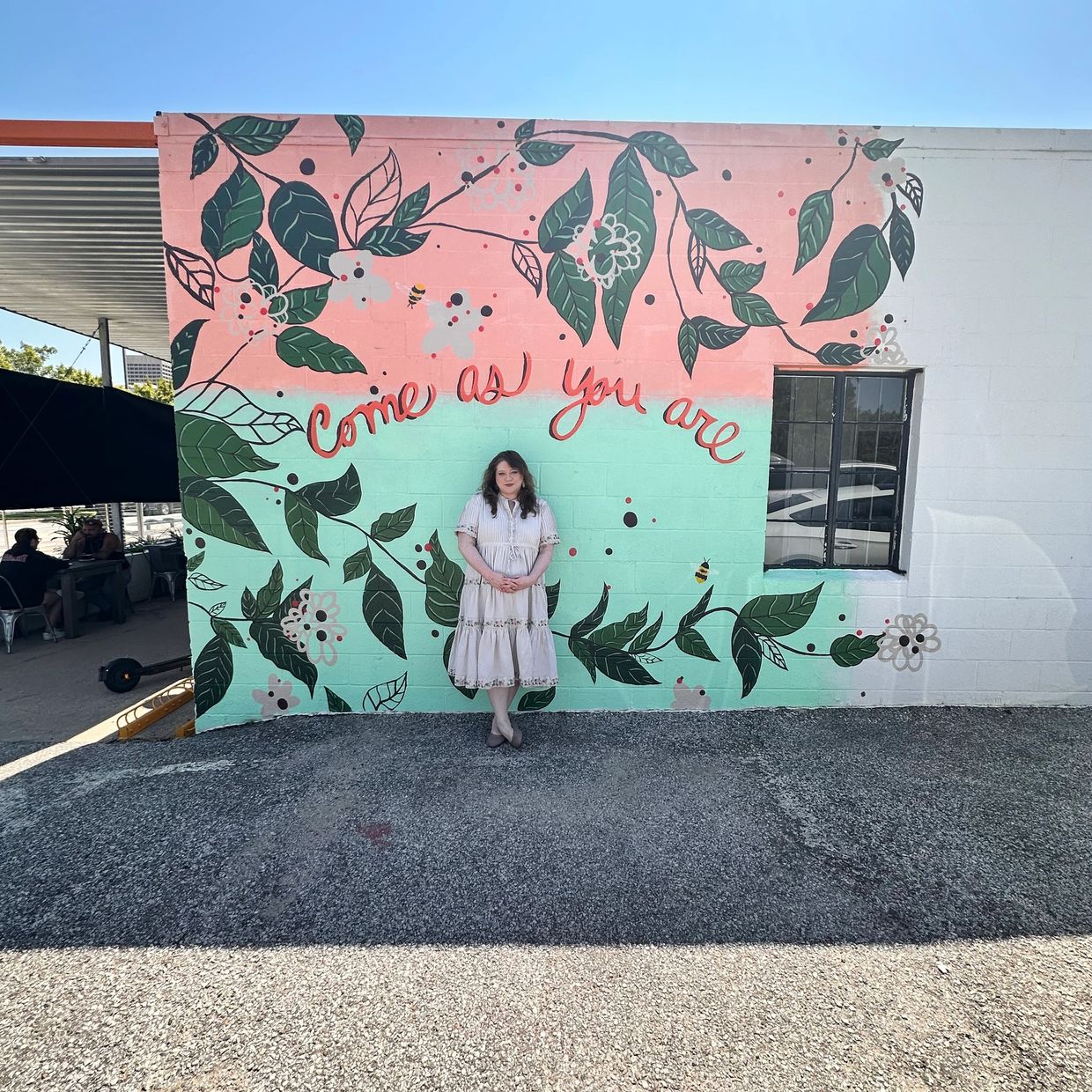 A woman stands in front of a colorful mural that says 'Come as you are'.