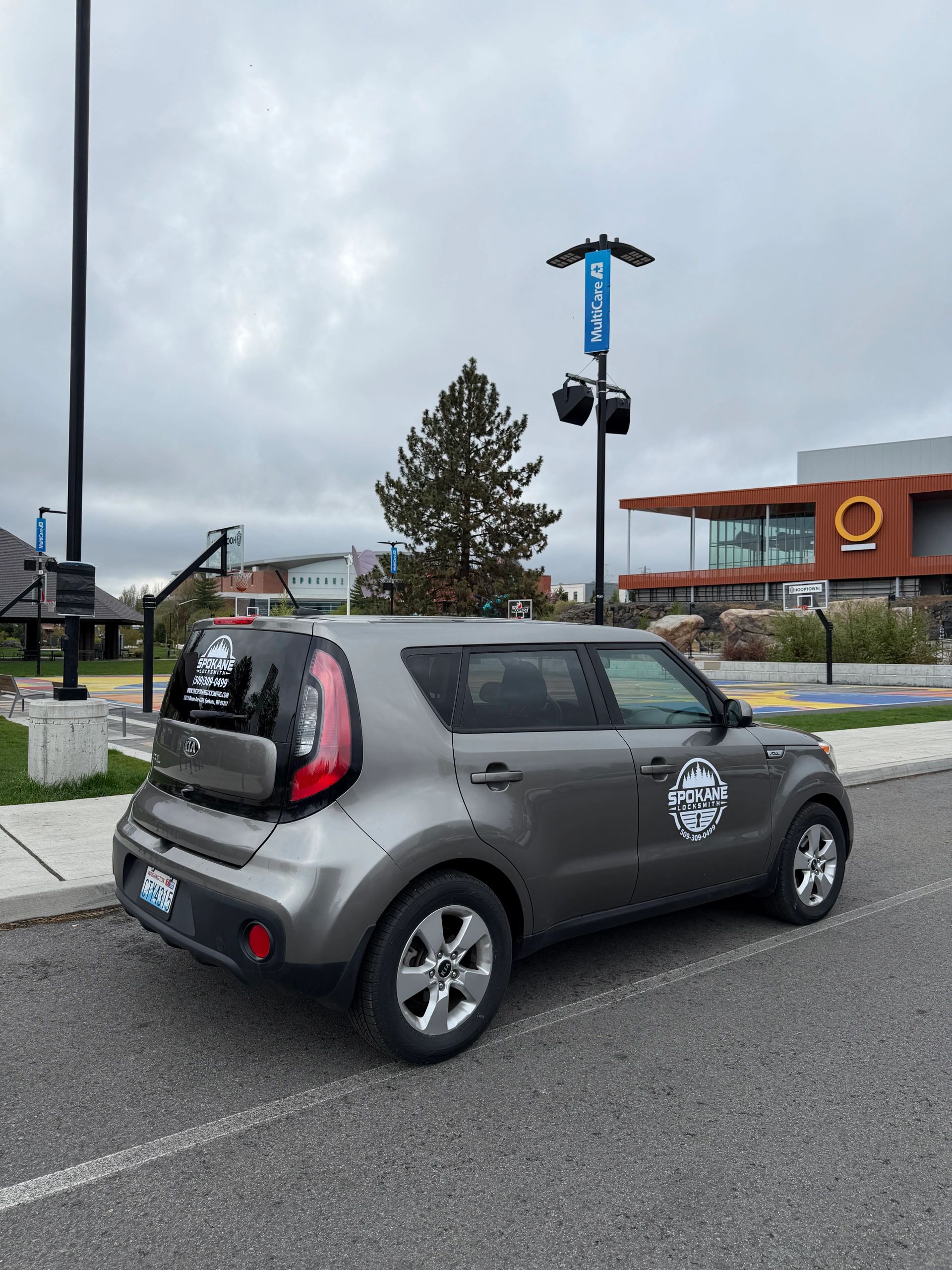 Gray Kia Soul with Spokane Locksmith logo parked near a basketball court under cloudy skies.