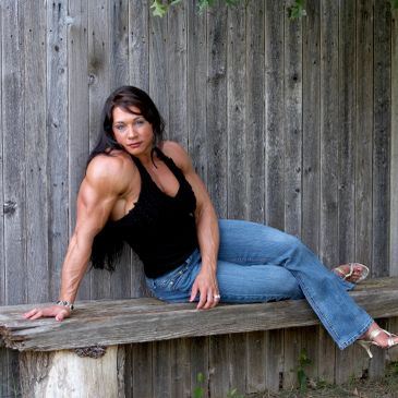 A muscular woman in jeans and heels poses against a rustic wooden fence.
