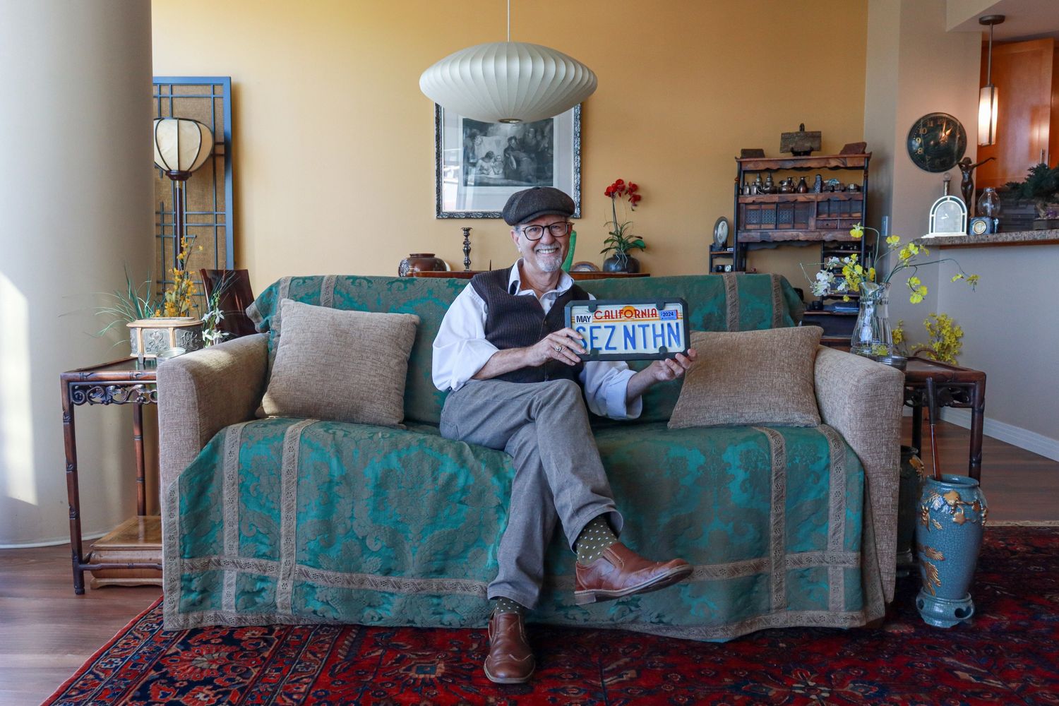 Elderly man smiling, sitting on a vintage sofa holding a personalized California license plate.