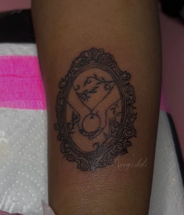 Ornate oval frame tattoo with a stylized hand holding a circle and floral elements inside.