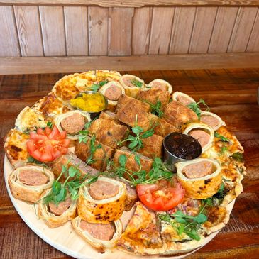 A plate with sausage rolls, pizza slices, fresh tomato, and dipping sauce.