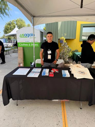 Man standing behind a table at a USAgain donation booth with informational materials and bags.