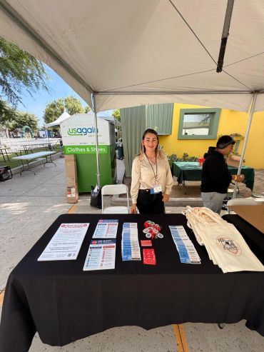 A woman stands behind a table with informational pamphlets and tote bags under a canopy tent.
