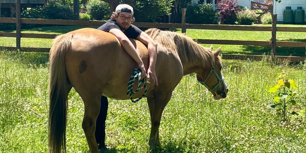 A man wearing a cap hugs a brown horse in a sunny grassy field.