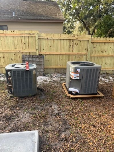 Two outdoor air conditioning units installed on the ground near a wooden fence.