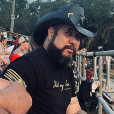 Man with a beard wearing a black cowboy hat and black t-shirt at an outdoor event.