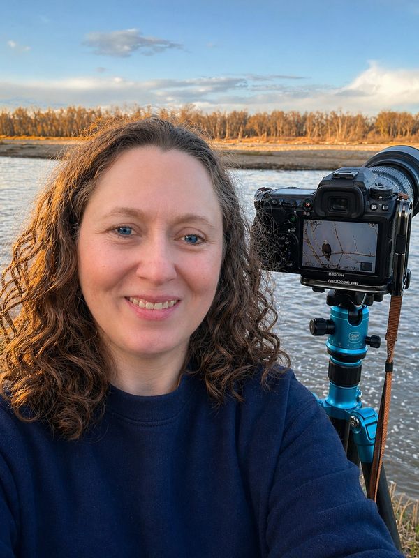Woman smiling near a river with a camera on a tripod.