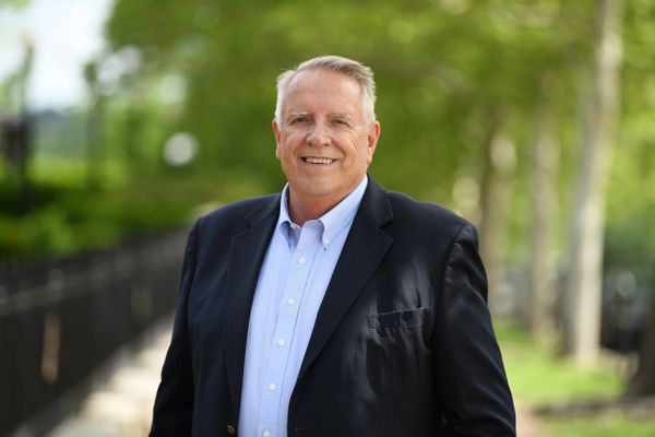Smiling older man in a blazer outdoors with trees in the background.