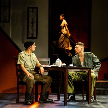 Two men in military uniforms sit at a table while a woman in a dress walks in the background.