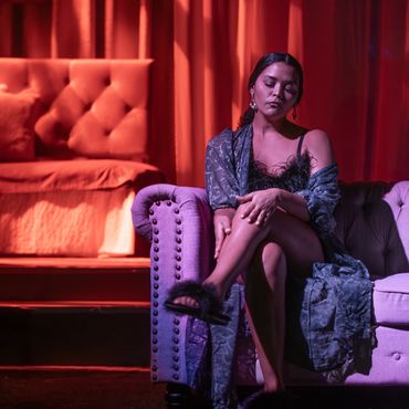 Woman in elegant attire sitting on a purple couch with red curtains in the background.