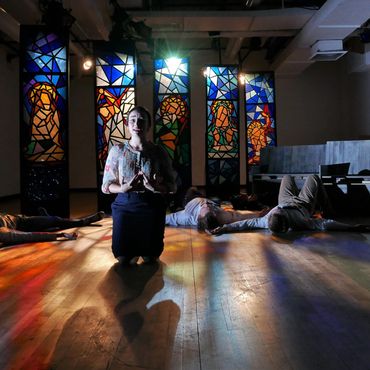 A woman kneels on stage with stained glass panels and people lying down behind her.