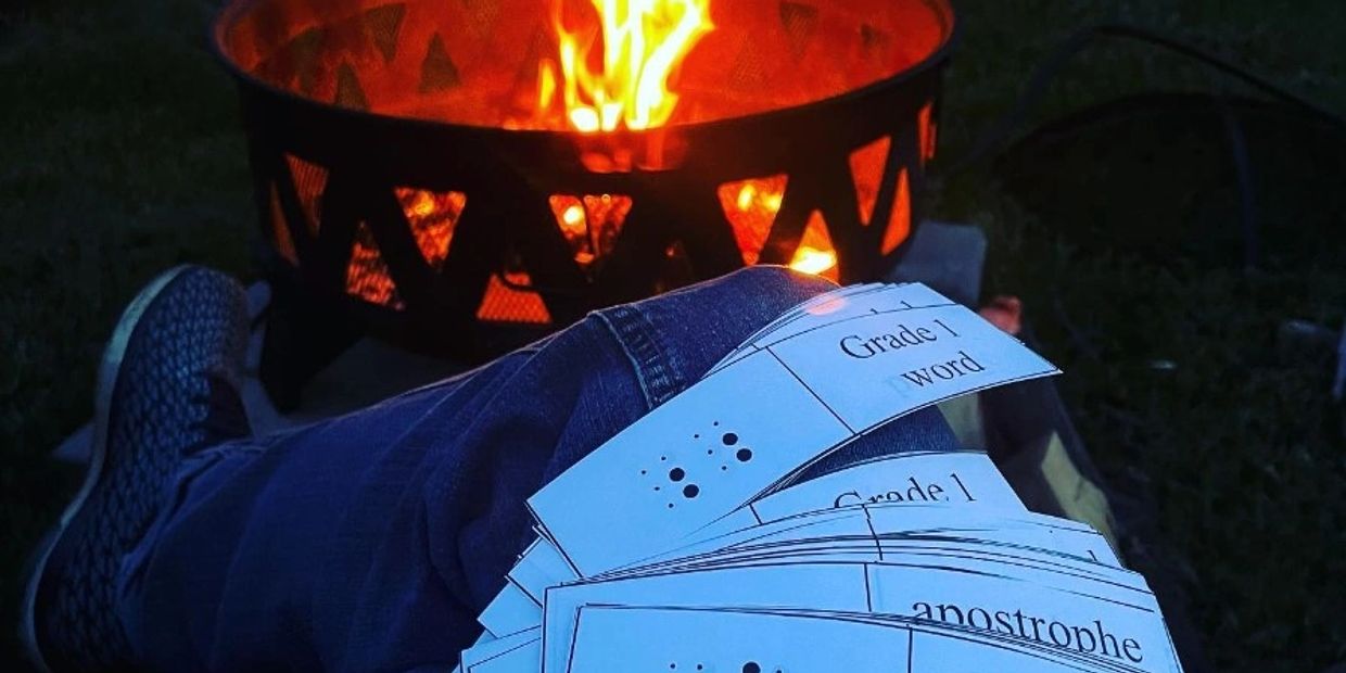 Elizabeth sitting by a campfire studying braille using SimBraille flashcards. 