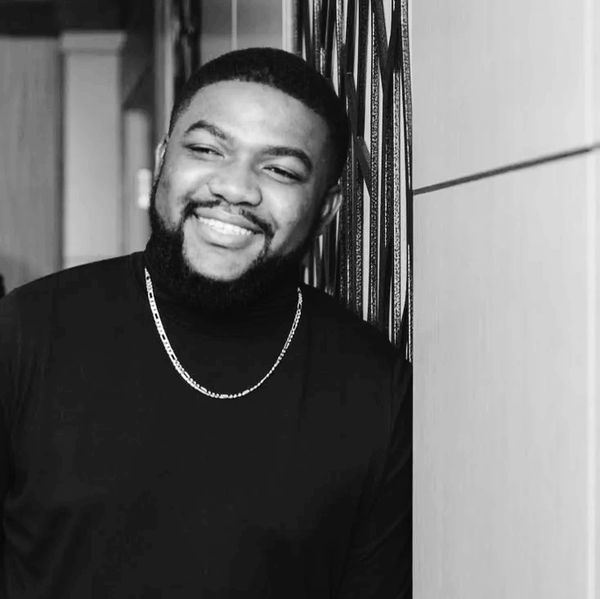 Smiling man in black turtleneck and chain leaning against a wall.