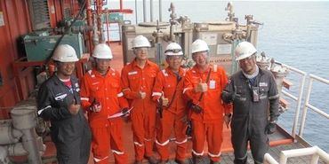 Group of six workers in safety gear giving thumbs up on an offshore platform.