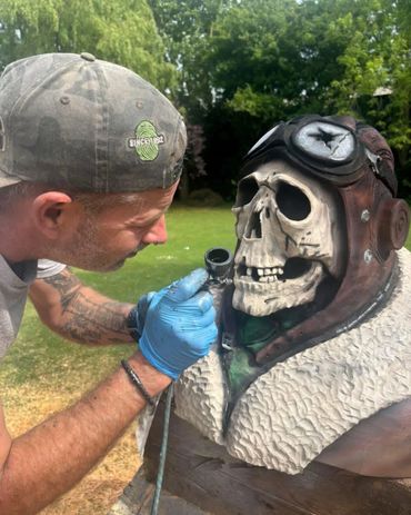 Man airbrushing details on a skull pilot sculpture outdoors.