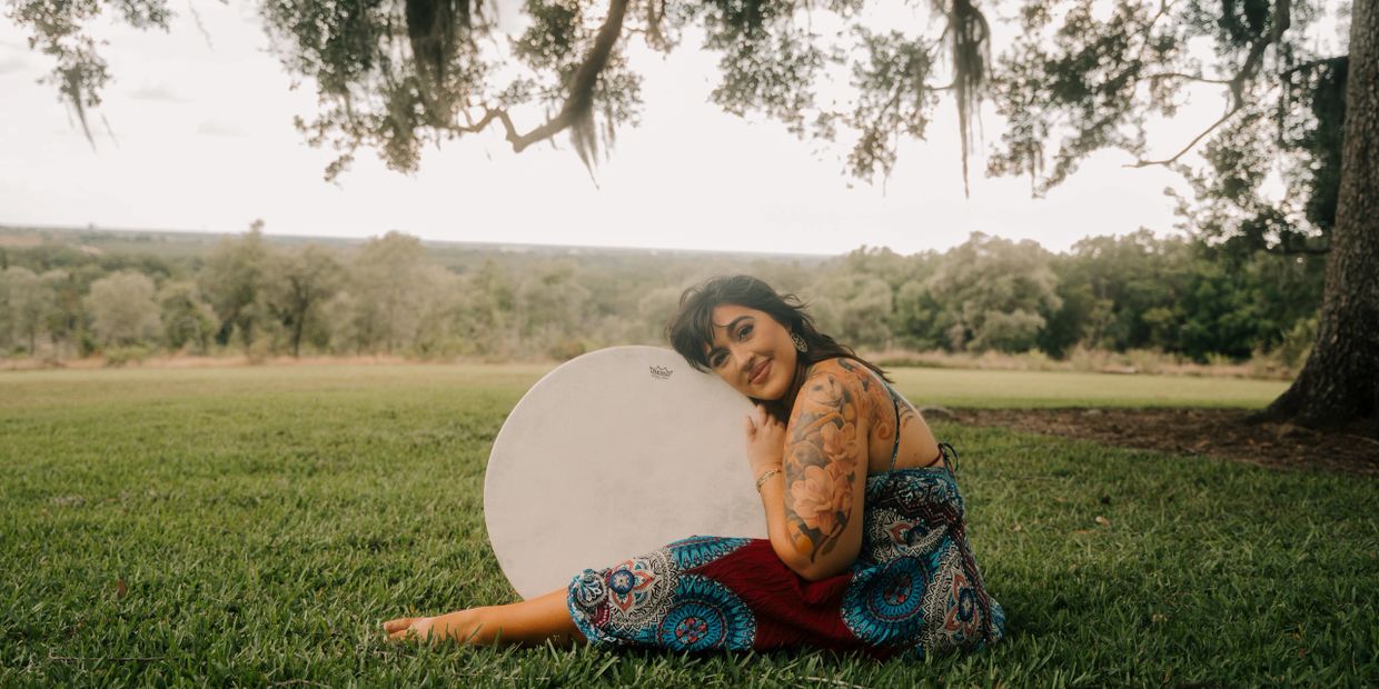 Woman with tattoos sits on grass hugging a drum outdoors.