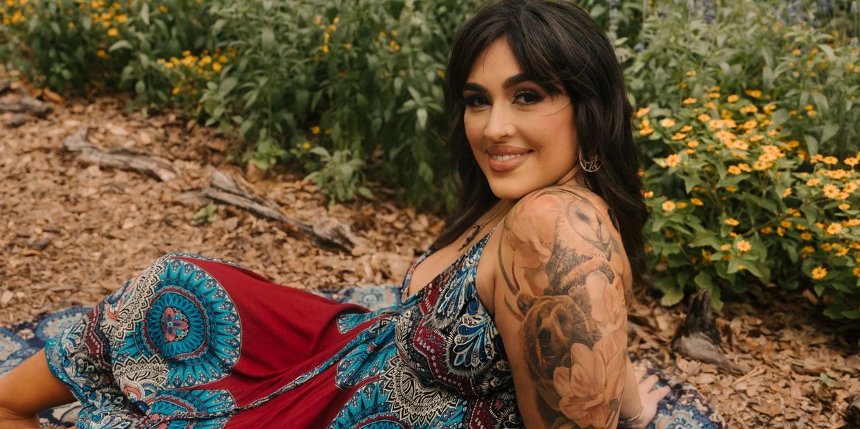 Woman with tattoo, smiling outdoors among flowers and wood chips.