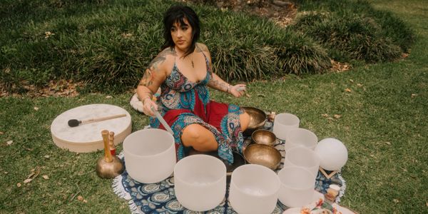Woman playing crystal singing bowls outdoors on a patterned cloth.