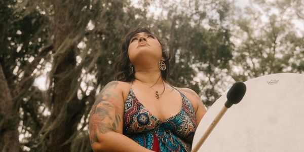 Woman with tattoos playing a drum in a natural outdoor setting.