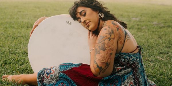 Woman with floral tattoos resting against a large drum outdoors.